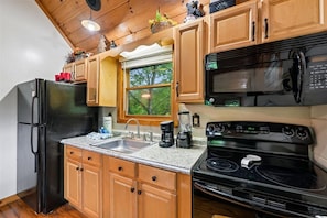 Une cuisine avec des meubles en bois, un réfrigérateur noir, une cuisinière, un four à micro-ondes et une fenêtre donnant sur des arbres.