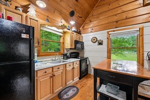 A kitchen with wooden cabinets, a black refrigerator, a stove, and a window overlooking greenery. - Serene Cozy Cabin w/ Hot Tub & Games (Sevierville)