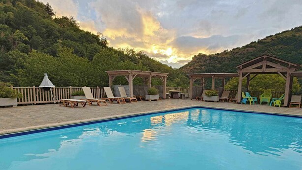 A swimming pool with lounge chairs and gazebos, surrounded by a wooden deck and a forested hillside.