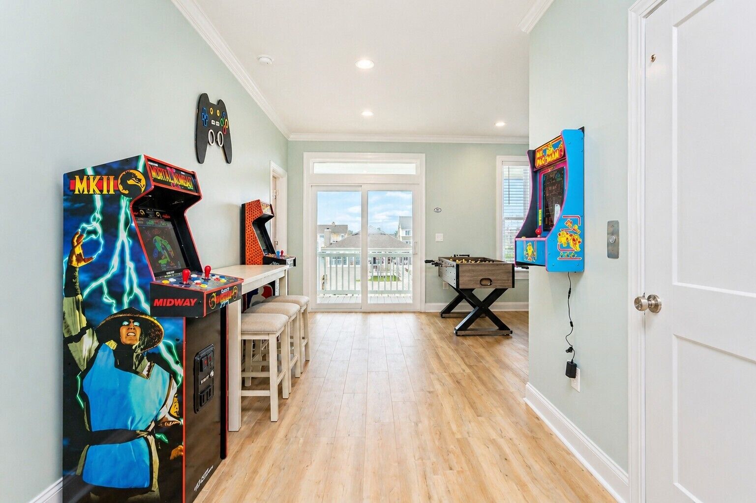 A room with a wooden floor, a foosball table, arcade games, and a view through a glass door.