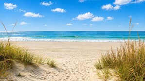 Een strand met zand en gras op de voorgrond, een heldere blauwe lucht en de oceaan in de verte.