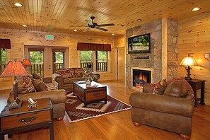 A log cabin interior with a fireplace, wooden furniture, and a ceiling fan. - Mountain Mansion w/ Views, Hot Tub & Games (Gatlinburg)