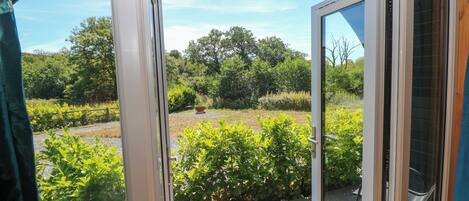 Uitzicht door een glazen deur op een houten terras met een tuin op de achtergrond.