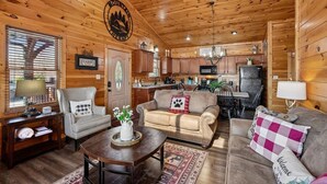 Ein Blockhaus-Interieur mit Holzbalkendecke, Essbereich, Küche und Wohnzimmer, möbliert mit Sofas und einem Couchtisch.