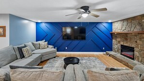 A modern living room with a sectional sofa, a flat-screen TV mounted on a blue wall, a fireplace, and a ceiling fan. - 4BR Home | Theater + Hot tub + Games + Fire Pit (Gatlinburg)