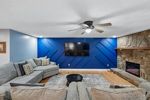 A modern living room with a sectional sofa, a flat-screen TV mounted on a blue wall, a fireplace, and a ceiling fan. - 4BR Home | Theater + Hot tub + Games + Fire Pit (Gatlinburg)