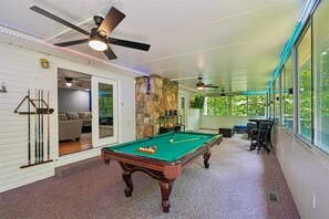 Ein Billardtisch in einem Raum mit Deckenventilator, einer Wand aus Stein und Blick durch große Fenster.