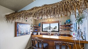 Una cocina con barra, sillas de madera, techo de paja y varios utensilios de cocina en estantes.