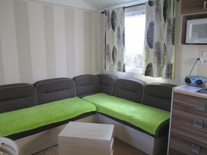 A compact room with a sofa, a small table, a cabinet, and curtains with a leaf pattern. - MOBILHOME 2 Bedrooms 4/5 pers camping 4* JULY AUGUST Rents 2025 idem to 2026 (Saint-Jean-de-Monts)