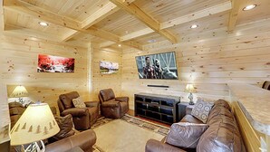 Interior acolhedor de uma cabana de toras, com lareira, assentos de couro e uma TV de tela plana na parede.