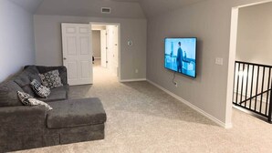 Una sala de estar con un sofá gris, un televisor de pantalla plana montado en la pared, un ventilador de techo y una puerta que da a otra habitación.