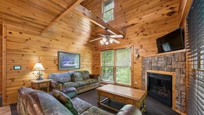L’intérieur d’une cabane en rondins, avec une cheminée en pierre, un ventilateur de plafond, un téléviseur et un tableau encadré au mur.