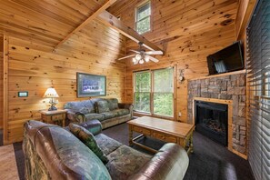 A log cabin interior with a stone fireplace, a ceiling fan, a television, and a framed picture on the wall. - Spacious Cabin w/ Game Room, Hot Tub & Fireplace (Gatlinburg)