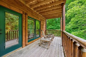 A wooden deck with chairs and a table, surrounded by green foliage. - Spacious Cabin w/ Game Room, Hot Tub & Fireplace (Gatlinburg)