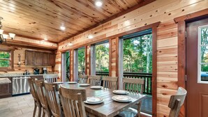 A wooden cabin interior with a dining area, kitchen, and large windows offering a view of the outdoors. - Glorious Mountain Lodge w/ Pool, Theater & Hot Tub (Gatlinburg)
