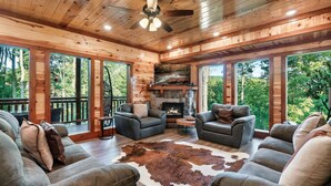 A wooden cabin interior with a stone fireplace, leather sofas, and large windows overlooking a forest. - Glorious Mountain Lodge w/ Pool, Theater & Hot Tub (Gatlinburg)