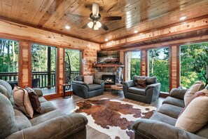 A wooden cabin interior with a stone fireplace, leather sofas, and large windows overlooking a forest. - Glorious Mountain Lodge w/ Pool, Theater & Hot Tub (Gatlinburg)