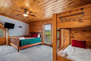 A wooden cabin room with a bed, a ceiling fan, a mounted TV, and a window with blinds. - Luxury Mountain Retreat w/ Hot Tub & Views (Pigeon Forge)