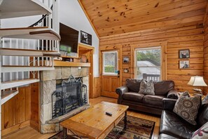 A cozy living room with a stone fireplace, wooden walls, a leather sofa, and a wooden coffee table. - Luxury Mountain Retreat w/ Hot Tub & Views (Pigeon Forge)