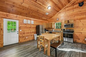 A wooden cabin interior with a dining area, kitchen, and a door leading to a green outdoor area. - Cozy Cabin w/ Fire Pit & Hot Tub (Pigeon Forge)