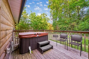 A wooden deck with a hot tub, two steps, and two wicker chairs. - Cozy Cabin w/ Fire Pit & Hot Tub (Pigeon Forge)