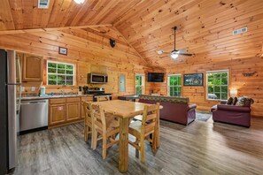 A cabin interior with a wooden ceiling, a dining area with a table and chairs, a kitchen with appliances, and a seating area with a couch and television. - Cozy Cabin w/ Fire Pit & Hot Tub (Pigeon Forge)