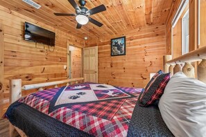 Ein Holzblock-Interieur mit einem Bett, einem Deckenventilator, einem an der Wand befestigten Fernseher und einer Tür.