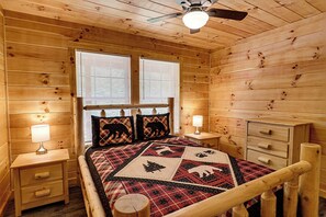Une chambre à coucher dans une cabane en bois, comprenant un lit, deux tables de chevet, une petite table, une commode et un ventilateur de plafond.
