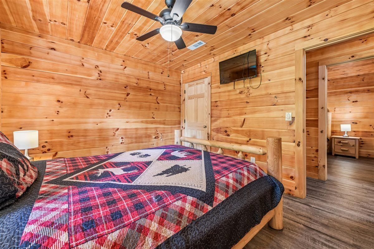 A wooden cabin room with a bed, a ceiling fan, a television, and a door leading to another room.