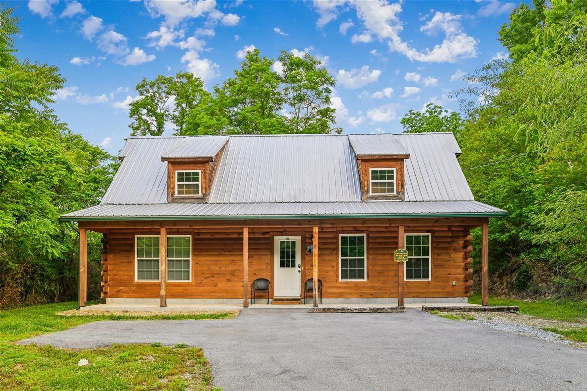 A log cabin with a metal roof, a front porch, and a gravel driveway.