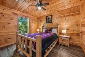 Un dormitorio de cabaña de madera con una cama, mesitas de noche, un ventilador de techo y una ventana.