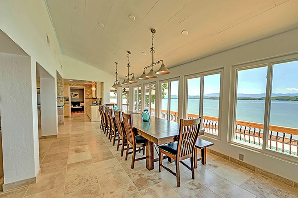 A long dining table with chairs, large windows with a view of the water, and a kitchen in the background.