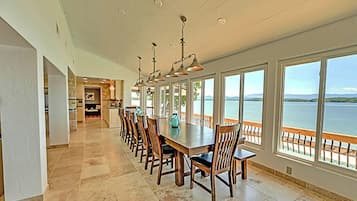 A long dining table with chairs, large windows with a view of the water, and a kitchen in the background.
