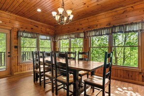 A wooden dining room with a large table and chairs, large windows with curtains, and a chandelier. - Chic Cabin w/ Theater & Fireplaces (Sevierville)