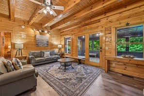 A log cabin interior with a fireplace, leather sofa, and a large window overlooking greenery. - Premier Riverfront Retreat w/ Hot Tub & Games (Pigeon Forge)