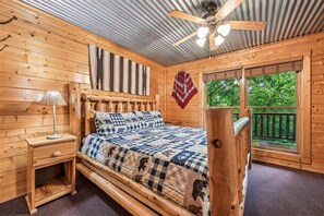 A wooden cabin bedroom with a bed, nightstand, lamp, ceiling fan, and a window with a view of trees. - Majestic Lodge w/ Views, Hot Tub & Games (Sevierville)