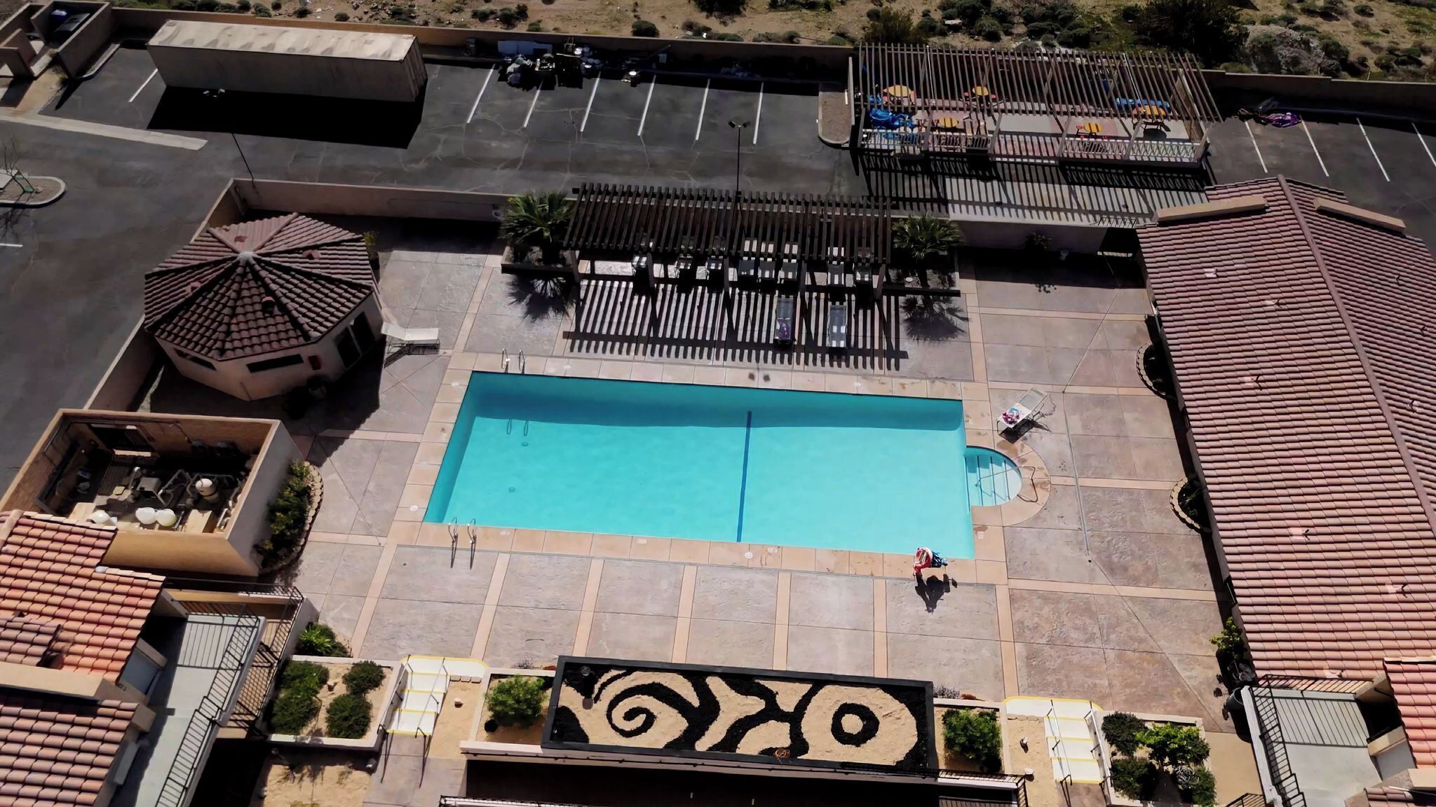 An aerial view of a resort with a central swimming pool, multiple lounge areas, and a parking lot.