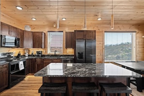 A modern kitchen with wooden cabinets, a large island with bar stools, stainless steel appliances, and large windows with blinds. - Modern Luxe Cabin w/ Views, Games & Hot Tub (Sevierville)