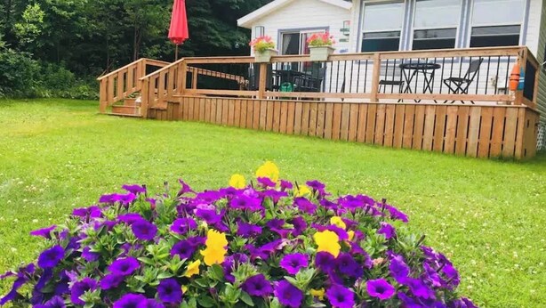 Une terrasse en bois avec une rambarde, un petit espace patio avec une table et des chaises, et un parterre de fleurs avec des fleurs violettes et jaunes.