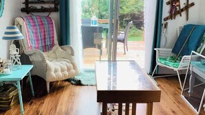 Un salon chaleureux avec une table en bois, un fauteuil rembourré et une chaise en osier. On y trouve une fenêtre avec des rideaux, une étagère avec des objets décoratifs et une vue sur l’extérieur.