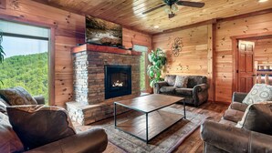 Interior acogedor de una cabaña de troncos con chimenea de piedra, sofás de cuero y un ventanal con vistas a un bosque.