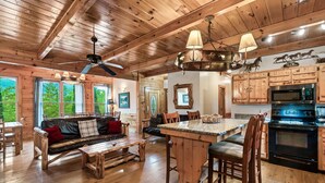 A wooden cabin interior with a dining area, sofa, kitchen, and large windows. - Grand Mountain Retreat w/ Games & Hot Tub (Pigeon Forge)
