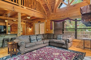 A cozy cabin interior with a wooden ceiling, a large sectional sofa, a bar area, and a view of the outdoors through large windows. - Mountain Cabin w/ Theater, Hot Tub & Views (Sevierville)