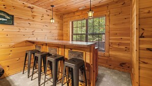 Interno di una baita in legno con angolo bar, sgabelli e una finestra vista sul verde.