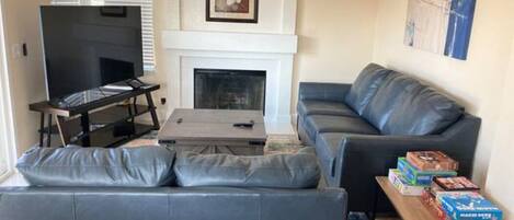 A living room with a grey sofa, a coffee table, a flat-screen TV, a fireplace, and a painting on the wall.