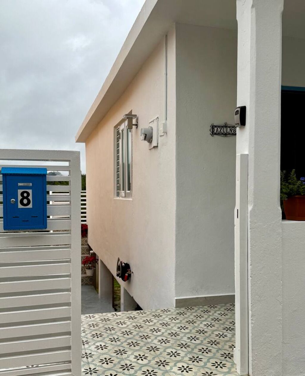 Ein schmaler Außenbereich mit einem blauen Briefkasten mit der Aufschrift '8', einem kleinen Fenster und einer Tür zu einem Zimmer.