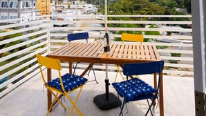 Ein Balkon mit Holztisch und Stühlen, einem beigen Sonnenschirm und Blick auf Gebäude und Grünflächen.