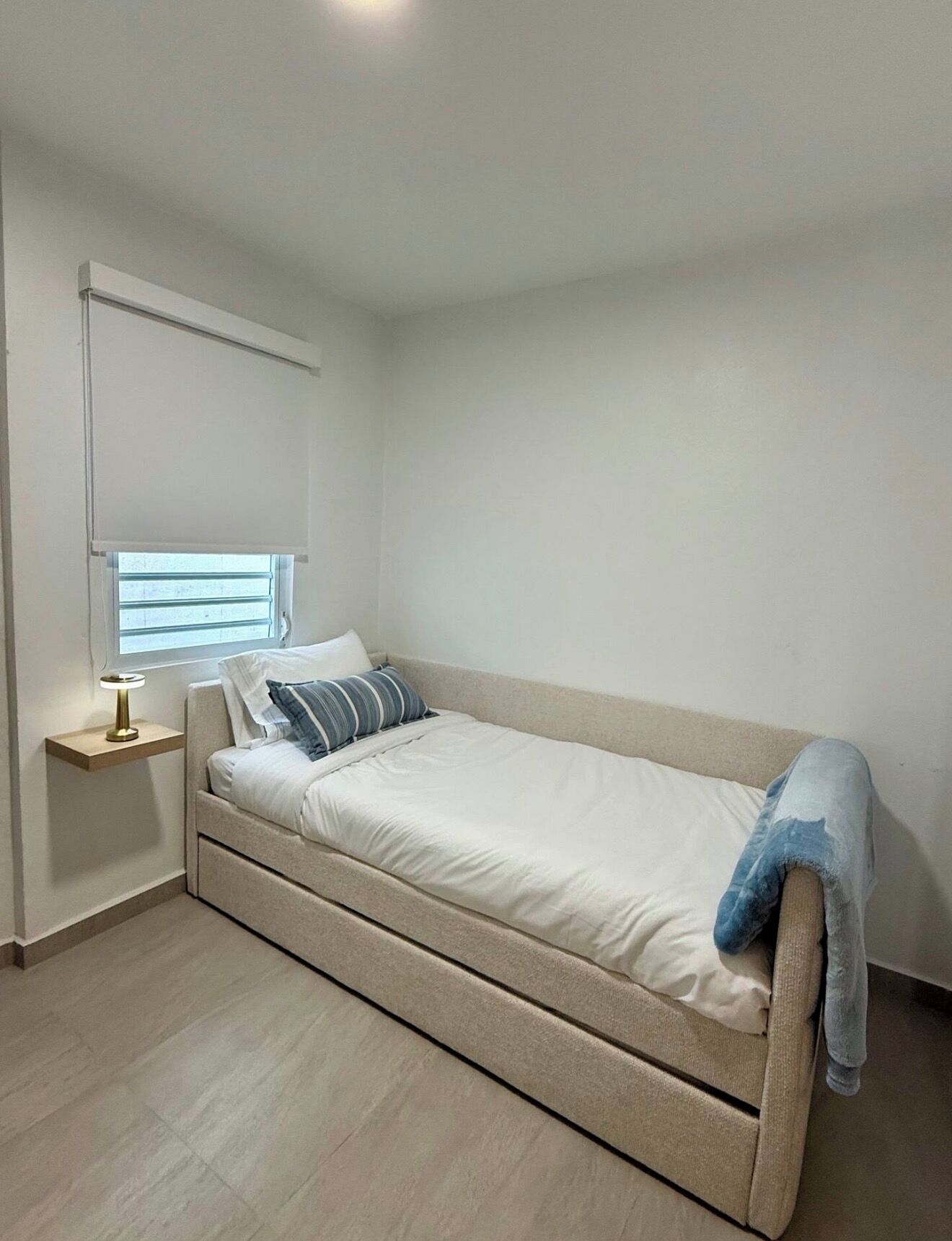 A single bed with a headboard, a bedside table with a lamp, and a window with blinds.