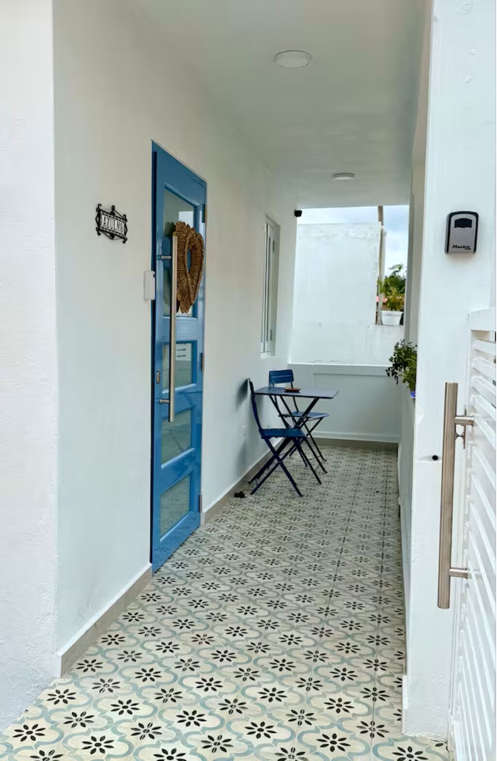 A narrow hallway with a blue door, a small table with chairs, and patterned floor tiles.