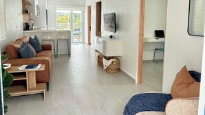 Ein modernes Wohnzimmer mit einer Couch, einem Couchtisch und einem Fernseher. Es gibt eine Küchenzeile mit Schränken und einem Fenster.